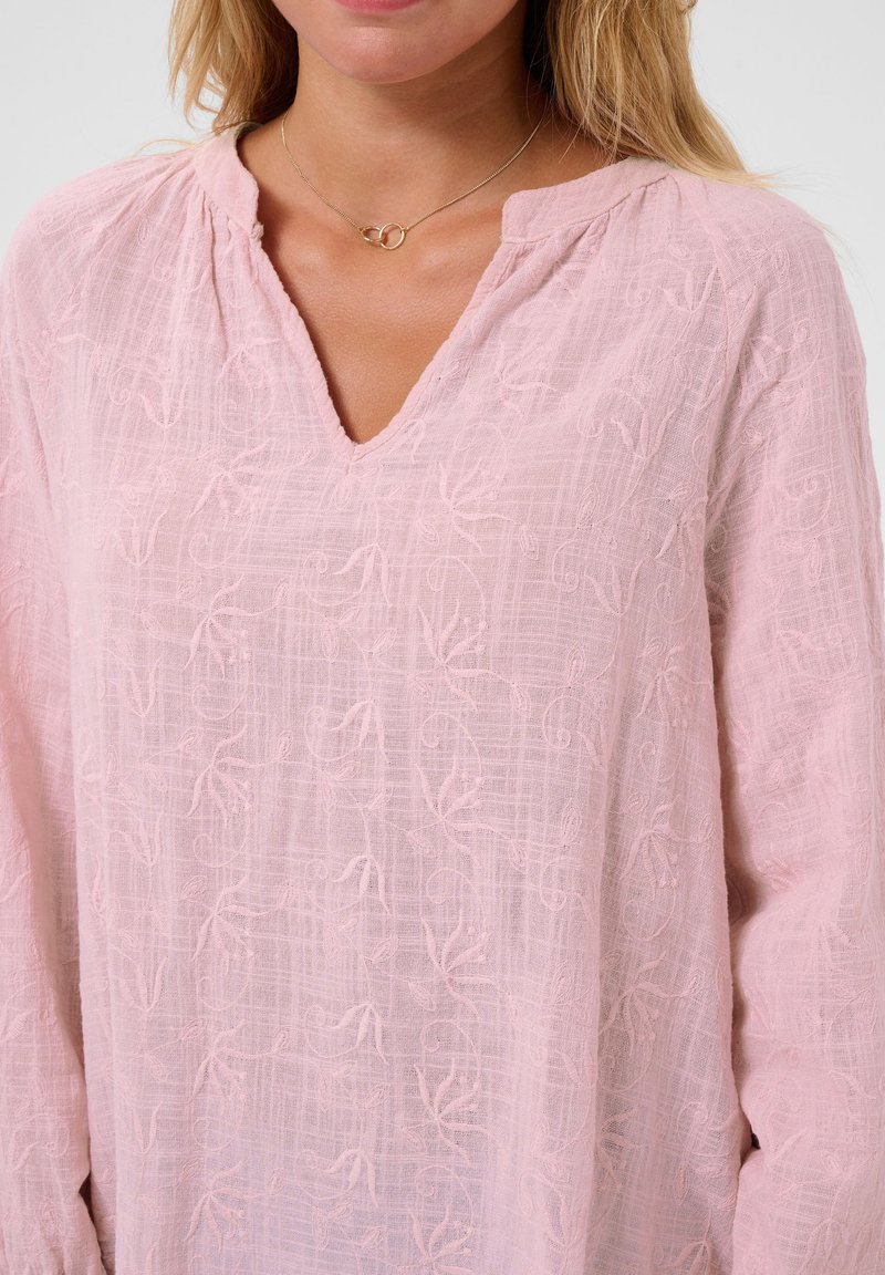 Femme portant un chemisier texturé rose clair avec un col en V et un délicat collier en or avec deux anneaux liés.