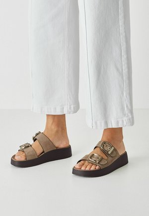 Sandales compensées en daim marron avec deux brides réglables, boucles en métal et semelle texturée foncée. Associées à un pantalon blanc à jambes larges.