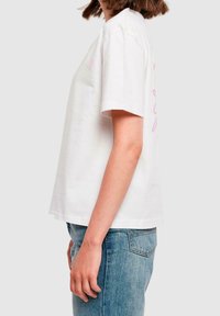 Bílá bavlněná tričko s krátkým rukávem, volný střih, s růžovým grafickým textem na boku, spárované se světle modrými džíny.