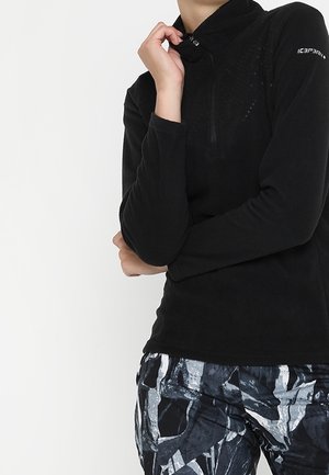 Person trägt ein schwarzes langärmeliges Oberteil mit Reißverschluss und abstrakt schwarz-weiß-grau gemusterte Hose, steht mit einer Hand in der Nähe des Kinns.