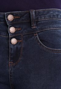 Tummansiniset farkut, joissa on korkea vyötärö, kolme kuparinväristä nappia, kontrastivärinen ompelu ja etupocket.