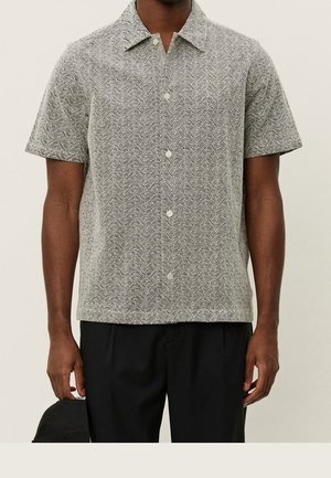 Mann trägt graues kurzärmeliges Hemd mit weißem Paisley-Muster und schwarze Hose, hält eine schwarze Kappe in der linken Hand.