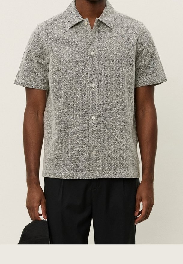 Homme portant une chemise grise à manches courtes avec un motif paisley blanc et un pantalon noir, tenant une casquette noire dans sa main gauche.
