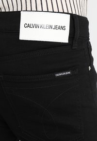 Svarta denimjeans med en vit läderlapp märkt "CALVIN KLEIN JEANS" på midjebandet. Har minimalistisk design och subtila sömdetaljer.