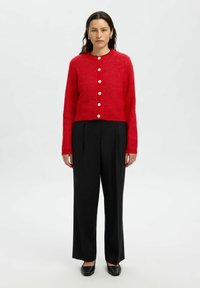 Kvinna som står mot en neutral bakgrund iförd en röd knappad cardigan, svarta vida byxor och svarta skor.