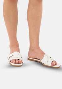 Sandalias deslizantes de cuero blanco con un diseño de tiras cruzadas, suela plana y sutiles acentos beiges. Dedos expuestos y estética minimalista.