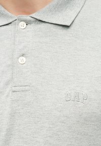 Ανοιχτό γκρι πόλο πουκάμισο με δύο λευκά κουμπιά και κεντημένο λογότυπο GAP στην αριστερή πλευρά του στήθους.