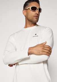 White long-sleeve shirt with a small black "snow peak" logo on the chest, paired with beige sunglasses. Smooth texture and relaxed fit.