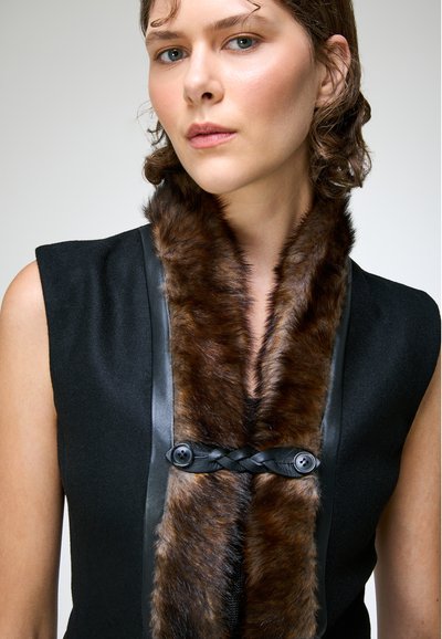 Femme portant un vêtement noir sans manches avec une fourrure brun foncé et un détail en cuir tressé noir avec des boutons au col.