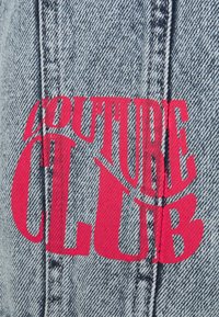 Denimtyg med en sliten textur, med en djärv röd "COUTURE CLUB" grafisk tryck framtill, mot en ljusblå bakgrund.
