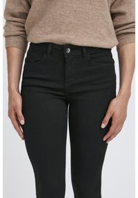 Jean en denim noir à taille haute, avec des poches avant et un détail de bouton argenté. Le tissu a une texture lisse et légèrement extensible.