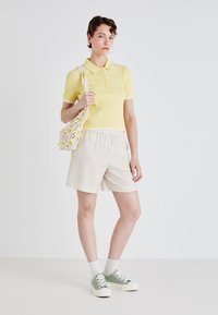 Gul ribbet polo skjorte med krage, beige shorts og grønne snørebåndssko. Tilbehør inkluderer en heklet veske med tusenfryd mønster.