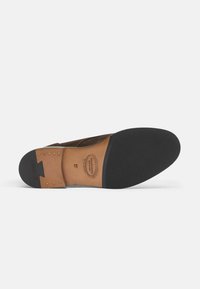 Suela de zapato de ante marrón con exterior de goma, inserción de cuero color beige, tachuelas metálicas y detalles de marca estampados. Diseño de punta redonda y plana.