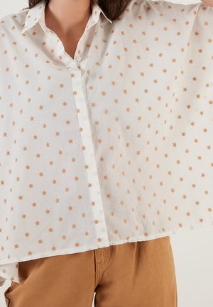 Vrouw draagt losse witte blouse met kleine bruine sterrenpatronen en lichtbruine broek, steekt haar arm op.