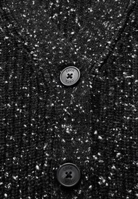 Czarny teksturowany kardigan z białymi kropkami, z dekoltem w serek i wyraźnym czarnym guzikiem z napisem "CECIL." Miękka, dzianinowa tkanina.