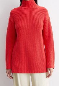 Femme portant un pull à col roulé en tricot côtelé rouge vif avec des manches longues et un pantalon crème, sur fond blanc.