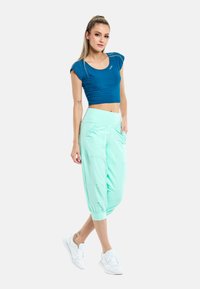 Top corto blu e pantaloni jogger verdi menta, tapered, con tasche laterali. Il tessuto appare liscio; la vita è ampia ed elastica. Scarpe da ginnastica bianche.