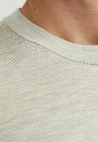 Světlý béžový pletený svetr zblízka, s texturovanou látkou, kulatým výstřihem a jemnými švy podél límce.