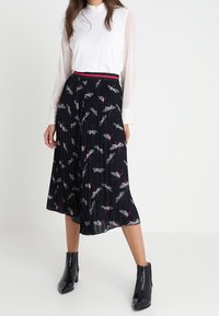 Gonna midi plissettata nera con motivo floreale colorato e cintura rossa, abbinata a una maglietta bianca e stivali neri alla caviglia.