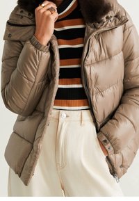 Kaki pufferjakke med en aftagelig pelskrave, lynlåslommer og trykknapper, lagd over en sort og orange stribet sweater.