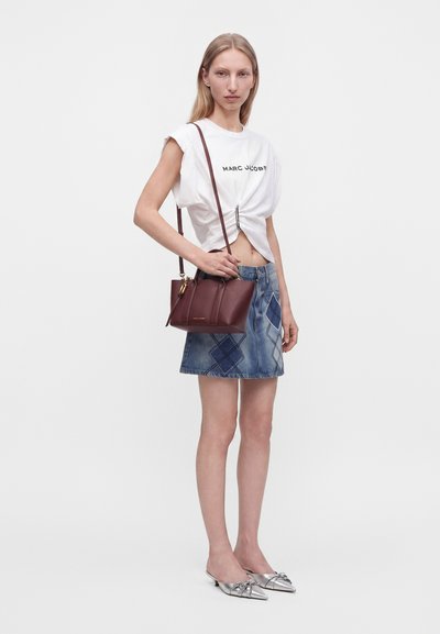 Νεαρή γυναίκα με λευκό μπλουζάκι Marc Jacobs, τζιν φούστα με διαμαντένια μπαλώματα, ασημένια μυτερά παπούτσια και κρατώντας καφέ δερμάτινη τσάντα ώμου.