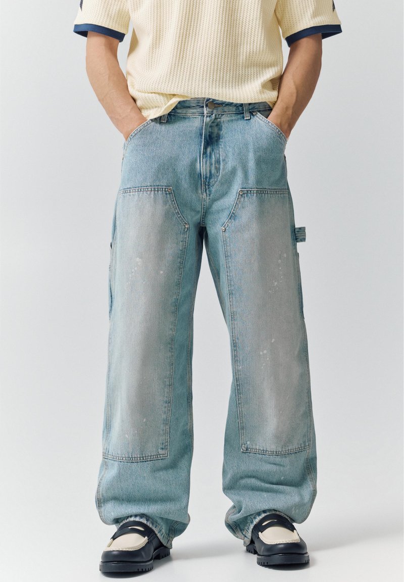 Osoba ubrana w luźne, wyblakłe niebieskie dżinsy z dużymi przednimi kieszeniami, kremowy krótki sweter z krótkim rękawem oraz czarne i białe masywne buty. Ręce w kieszeniach.