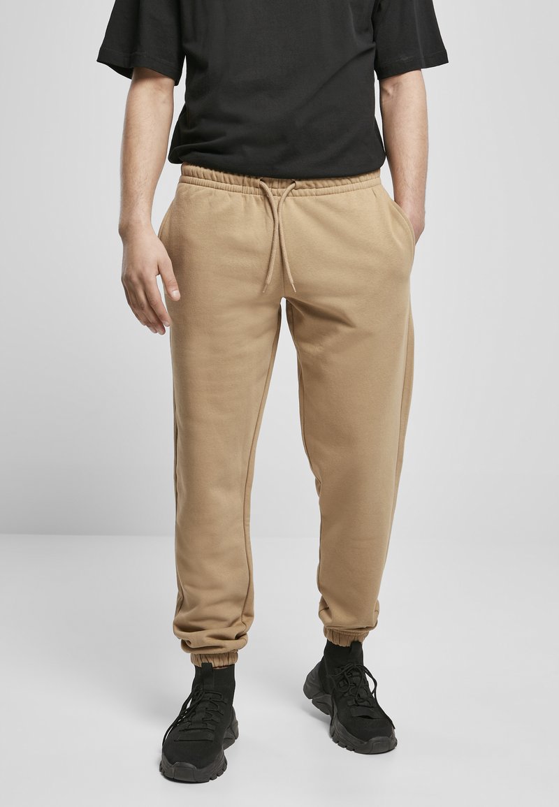 Beige joggebukser med elastisk midje og mansjetter, med sidelommer og snøring, sammen med svarte treningssko.