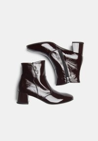 Botas de tobillo de un marrón oscuro brillante con tacones de bloque, cremalleras laterales y sutiles acentos metálicos decorativos en una de las botas, sobre un fondo blanco.