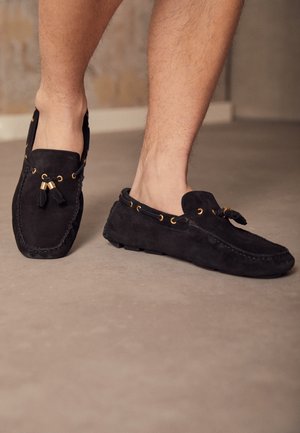 Musta mokkanahkainen loafer-kenkä, jossa on kullanväriset silmukat ja hapsut, jalassa paljain jaloin, seisomassa sileällä beigenvärisellä lattialla sisätiloissa.