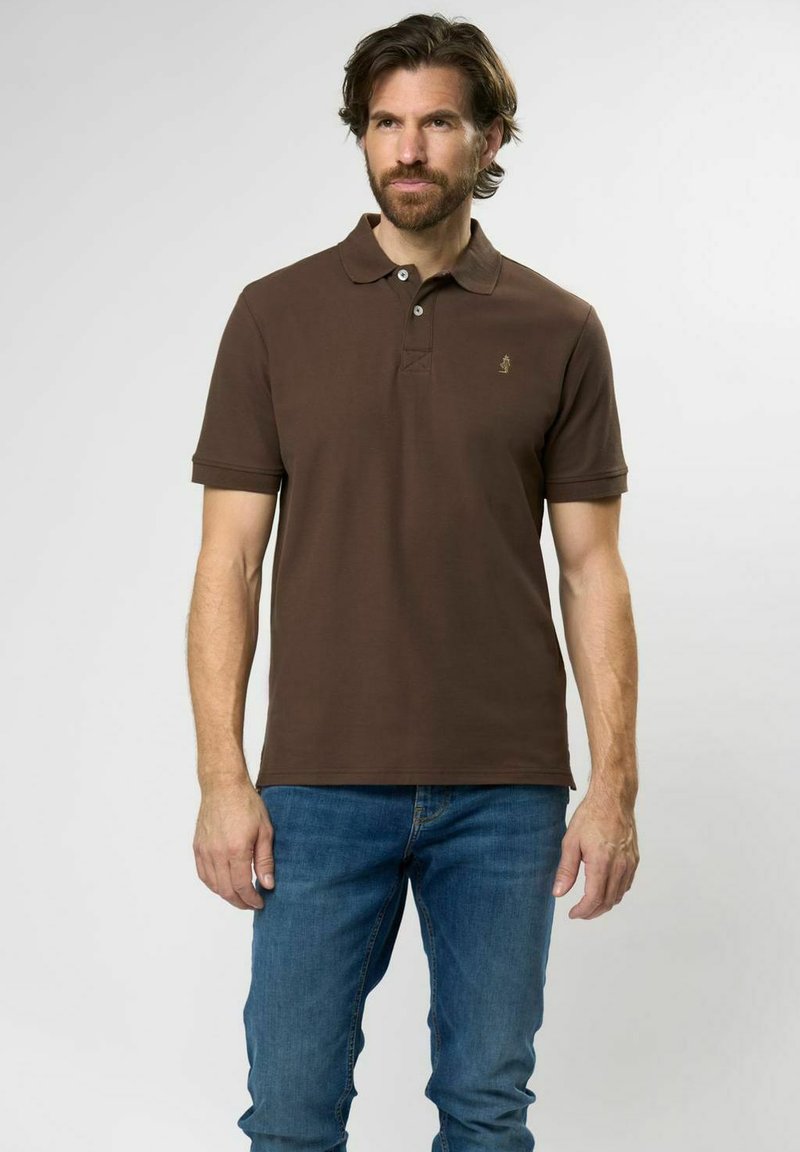 Braunes Poloshirt mit Kragen und drei Knöpfen, mit kurzen Ärmeln und einem kleinen Logo auf der linken Brust. Kombiniert mit blauen Jeans.