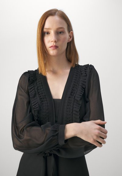 Blouse noire avec des détails à volants sur le devant, des manches longues transparentes et un décolleté en V. Texture lisse avec une silhouette légèrement ajustée.