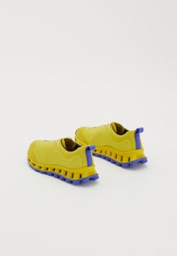 Tênis de desporto amarelos, tricotados, com solas e partes traseiras azuis, exibidos lado a lado em fundo branco.