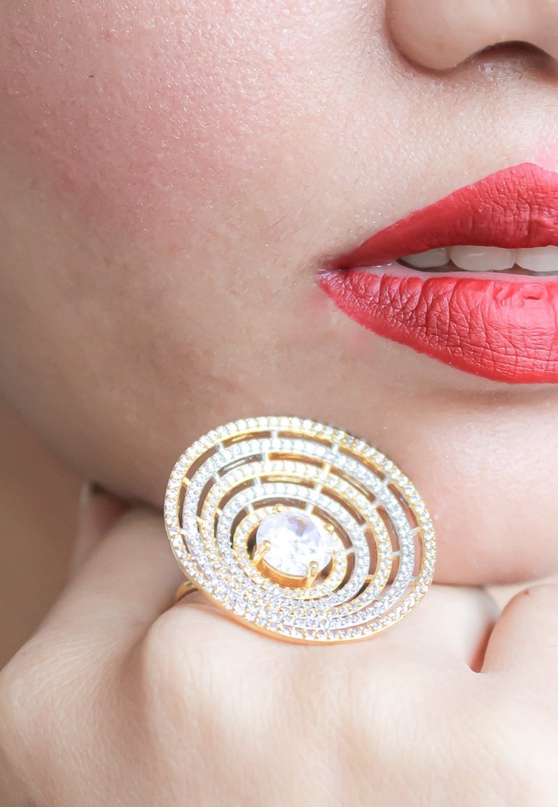 Gros plan d'une main portant une grande bague circulaire en or et diamants près des lèvres avec un rouge à lèvres rouge vif.