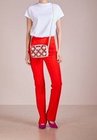 Red tailored pants, white t-shirt, and a patterned crossbody bag with pink accents. Model wears purple heels. Simple background.