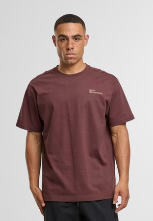 Ein Mann mit kurzhaariger Frisur und Sommersprossen trägt ein schlichtes dunkelrotes T-Shirt mit kleinem, hellem Text auf der linken Brust und steht vor einem weißen Hintergrund.