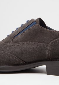 Chaussure en daim gris avec des accents en tissu bleu, présentant des coutures détaillées et un petit talon bloc. Les côtés ont une forme arrondie avec des courbes subtiles.
