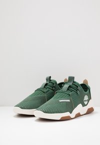 Baskets tricotées vertes avec une finition texturée, présentant un logo blanc, une semelle en caoutchouc et des tirettes. Accentuées de détails marron clair.