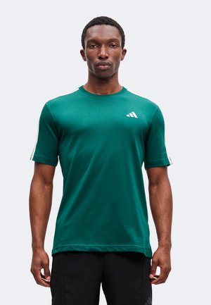 Άνδρας που φοράει πράσινο μπλουζάκι Adidas με λευκές ρίγες στα μανίκια και μαύρο παντελόνι, στέκεται μπροστά σε απλό φωτεινό φόντο.