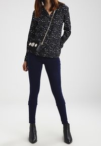 Marineblå bluse med lange ærmer og et prikket mønster, parret med mørke skinny jeans og sorte spidse ankelstøvler, mens der holdes en lille pyntet taske.