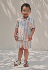 Jeune garçon debout en chemise rayée à manches courtes et short assorti avec des sandales grises à boucle sur un fond clair et ombragé.