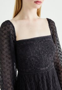 Czarna sukienka wyszywana, z długimi przeźroczystymi rękawami, ozdobiona skomplikowanymi wzorami kwiatowymi oraz kwadratowym dekoltem, o teksturowanej tkaninie i delikatnych koronkowych akcentach.