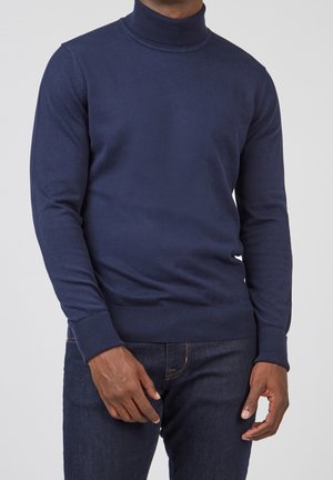 Mann trägt einen enganliegenden dunkelblauen Rollkragenpullover und dunkle blaue Jeans, steht vor einem schlichten hellen Hintergrund.