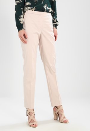 Femme portant un pantalon beige clair sur mesure, un chemisier vert à fleurs et des sandales beiges à talons hauts avec des brides croisées, debout sur un sol blanc.