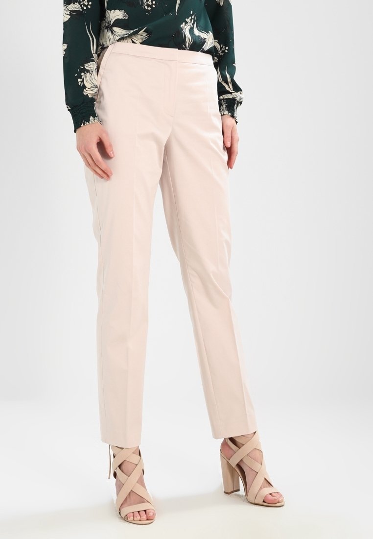 Femme portant un pantalon beige clair sur mesure, un chemisier vert à fleurs et des sandales beiges à talons hauts avec des brides croisées, debout sur un sol blanc.