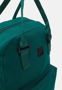 Un sac à dos vert en tissu durable avec une texture lisse, comprenant deux poignées, des compartiments zippés et un patch de logo noir.