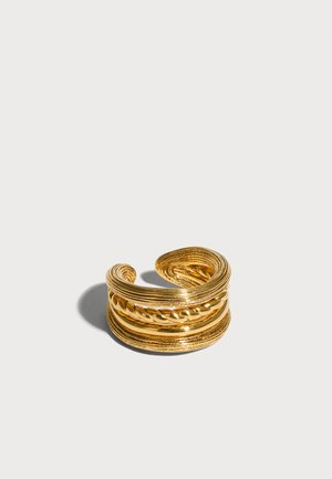 Åben guldfarvet manchet ring med lagdelte og snoede teksturerede bånd mod en lysegrå baggrund.