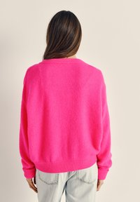 Persona con capelli castani lunghi che indossa un maglione rosa acceso e jeans azzurri chiari, vista di spalle davanti a uno sfondo beige semplice.