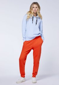 Helltürkisfarbener Kapuzenpullover mit schwarzen Kordeln, Fronttasche, kombiniert mit orangen Jogginghosen mit einem Logo nahe dem Saum und weißen Turnschuhen.