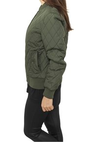 Una persona in una giacca bomber verde oliva trapuntata, in profilo, abbinata a leggings neri, con capelli lunghi parzialmente visibili.