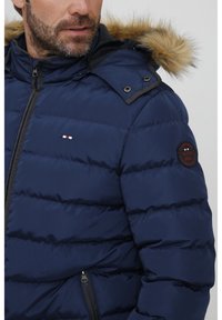 Marineblå puffjakke med pelsforet hette, quiltet tekstur, glidelås, og logo-patch på ermet. Har trykk- og glidelåslommer.